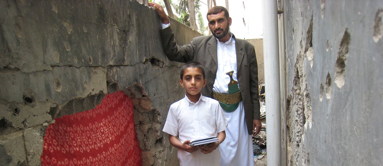 Een jongen en een man staan in steegje naast een beschadigde muur. Hun huis werd beschoten terwijl de hele familie in een van de kamers schuilde (juli 2012).