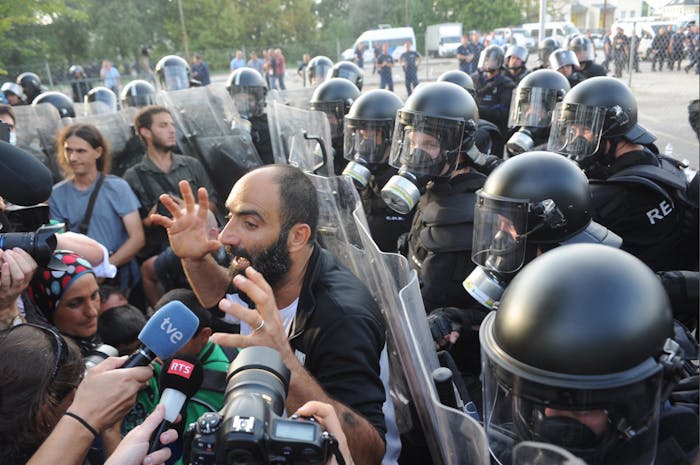 Ahmed H. praat in 2015 bij de grens tussen Servië en Hongarije met de media, met achter hem de Hongaarse oproerpolitie. De politie zette traangas en een waterkanon in om migranten te verhinderen het land binnen te komen.