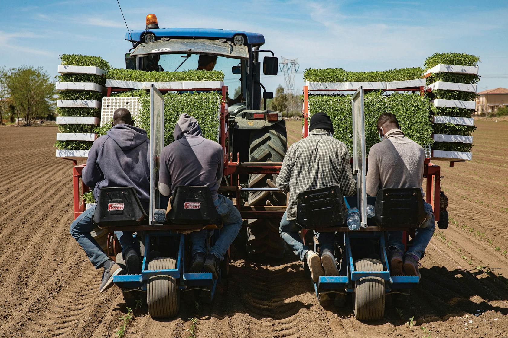 Seizoensarbeiders op een tractor. Tussen hen in een paneel tegen de verspreiding van het virus.