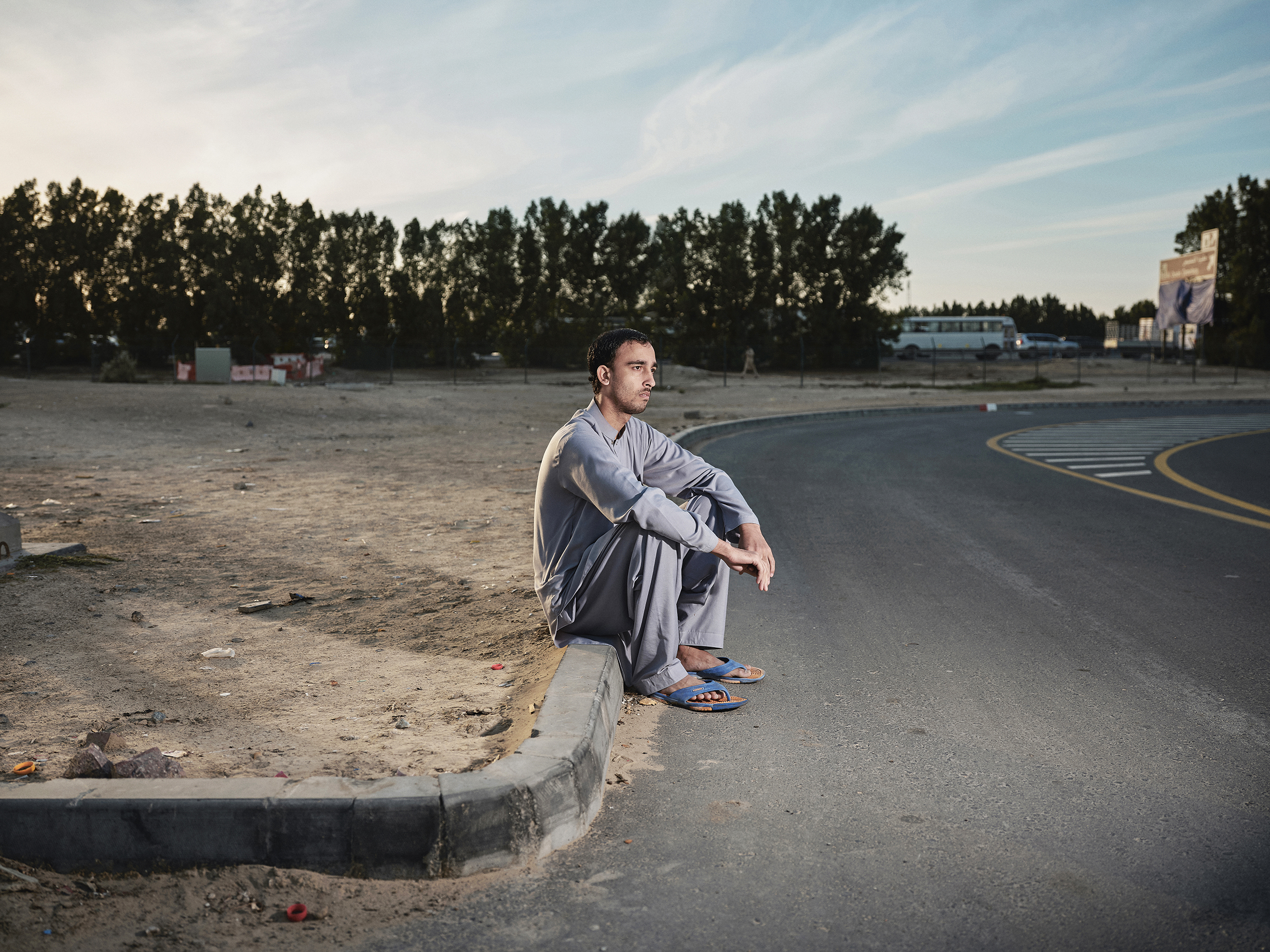 Een man zit op straat vlak bij het werkkamp waar hij woont. Veel arbeidsmigranten wonen in dit soort kampen: dicht op elkaar en met weinig sanitaire voorzieningen.