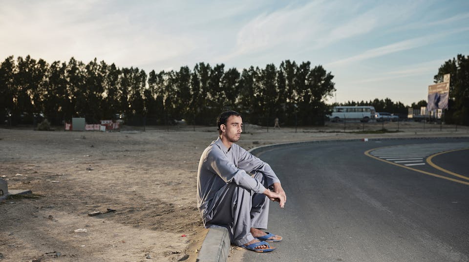 Een man zit op straat vlak bij het werkkamp waar hij woont. Veel arbeidsmigranten wonen in dit soort kampen: dicht op elkaar en met weinig sanitaire voorzieningen.