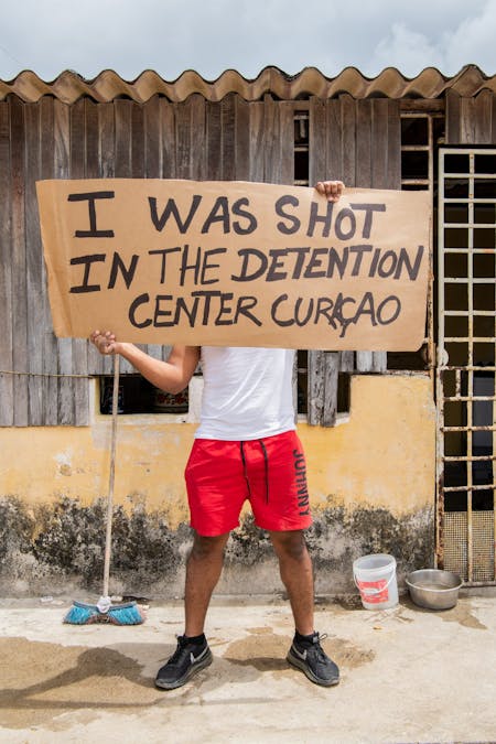 Een Venezolaanse man die illegaal op Curaçao woont houdt een stuk karton vast met daarop de tekst: Ik werd neergeschoten in het detentiecentrum Curaçao.