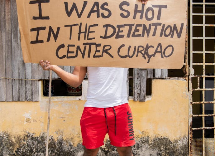 Een Venezolaanse man die illegaal op Curaçao woont houdt een stuk karton vast met daarop de tekst: Ik werd neergeschoten in het detentiecentrum Curaçao.