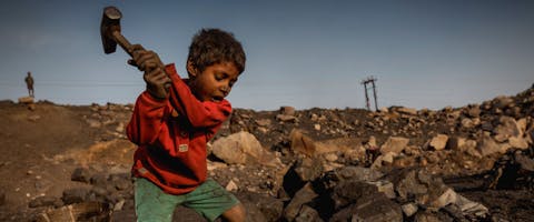 Een kleine jongen is aan het werk in een kolenveld in Jharia. Hij draagt een rode trui en een groene korte broek.