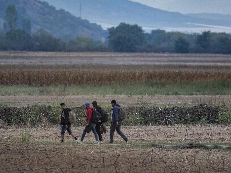 Een groep vluchtelingen en migranten, voornamelijk mannen uit Afghanistan, loopt in de velden aan de Griekse kant met het Noord-Macedonische gebergte op de achtergrond. Ze proberen de Grieks-Noord-Macedonische grens over te steken om de Balkanroute te volgen, die in 2015-2016 beroemd werd tijdens de Syrische vluchtelingencrisis, van Idomeni in Griekenland naar Gevgelija in Noord-Macedonië, waarbij ze de treinrails en het treinstation volgen, om vervolgens Midden- en Noord-Europa te bereiken. Ze staan voor een hek dat de twee landen scheidt. De foto is genomen op 6 oktober 2021 en toont de vluchtelingen en migranten die de grenzen proberen te passeren.