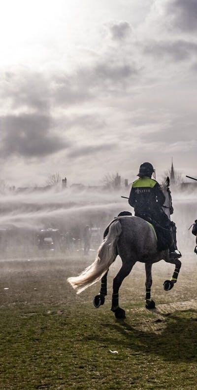 Politie zet waterkanonnen in tijdens protest tegen corona maatregelen. Je ziet 2 politie op paarden met hun rug naar de camera gekeerd. Er worden waterkanonnen ingezet.