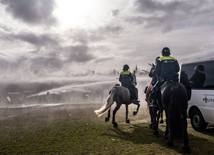Politie zet waterkanonnen in tijdens protest tegen corona maatregelen. Je ziet 2 politie op paarden met hun rug naar de camera gekeerd. Er worden waterkanonnen ingezet.