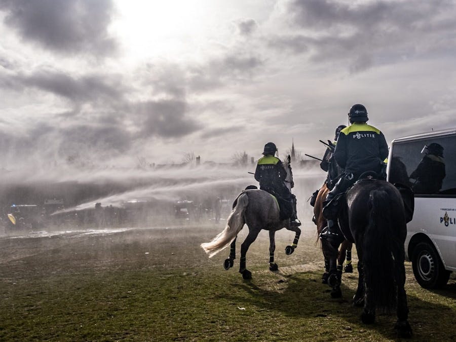 Politie zet waterkanonnen in tijdens protest tegen corona maatregelen. Je ziet 2 politie op paarden met hun rug naar de camera gekeerd. Er worden waterkanonnen ingezet.
