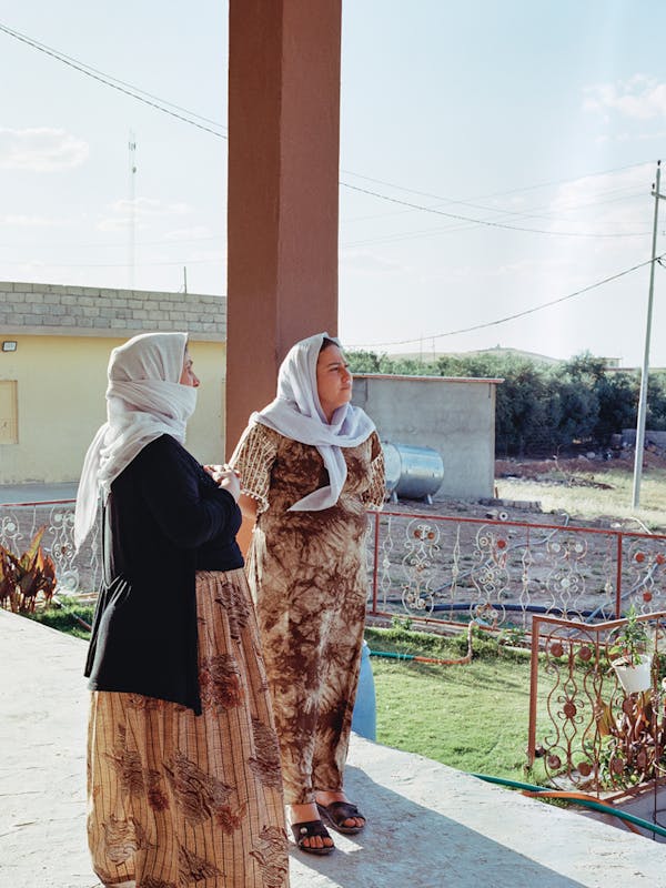 Twee vrouwen staan voor hun huis in Sherfedin, een plaats met een van de van grootste Jezidi-heiligdommen in het gebied. Door meerdere wonderen zou het behouden zijn gebleven in de IS-tijd.